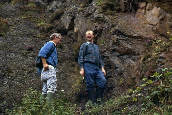 Two men standing next to a wall of rock.