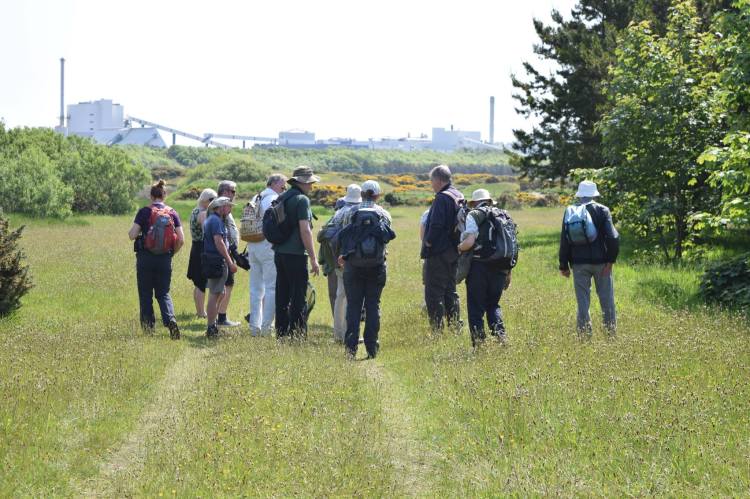Group of 12 members standing in open grassland with industrial buildings in background.