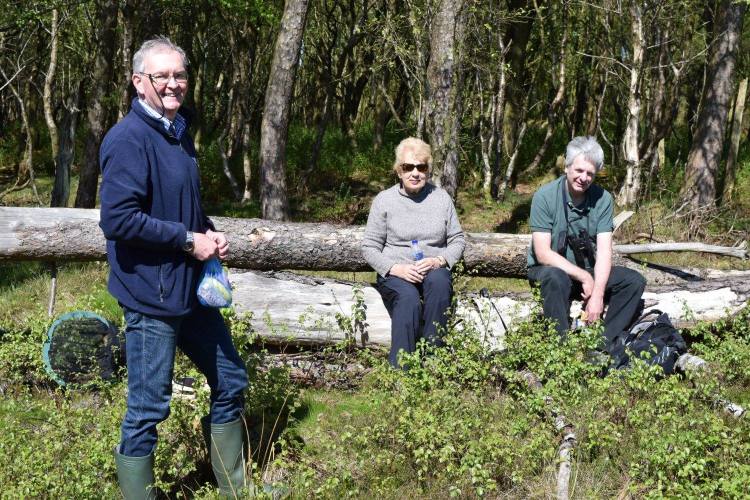 Two members sitting on felled trees beside woodland and one person standing all with pack lunches in hand.
