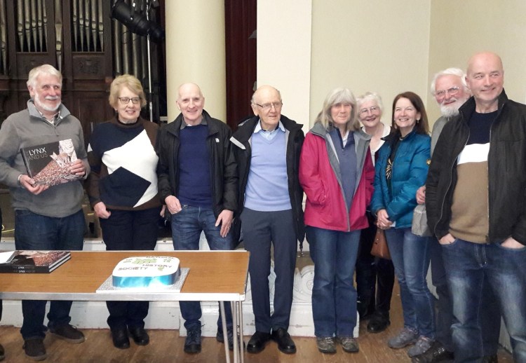 Group of nine members standing in a row behind a table with a 50th anniversary cake on it.