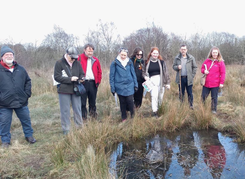 Group of 8 people standing next to a pond full of frogs with rough grassland and trees behind
