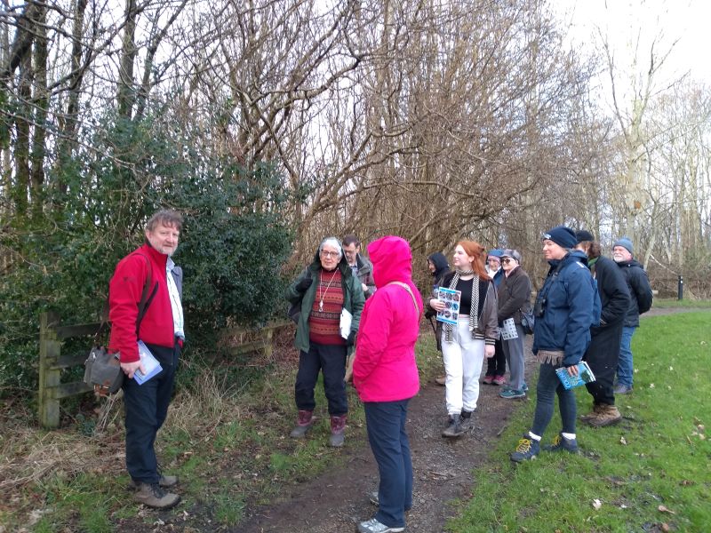 Group of 11 people standing around a path next to a holly bush with mature leafless trees behind