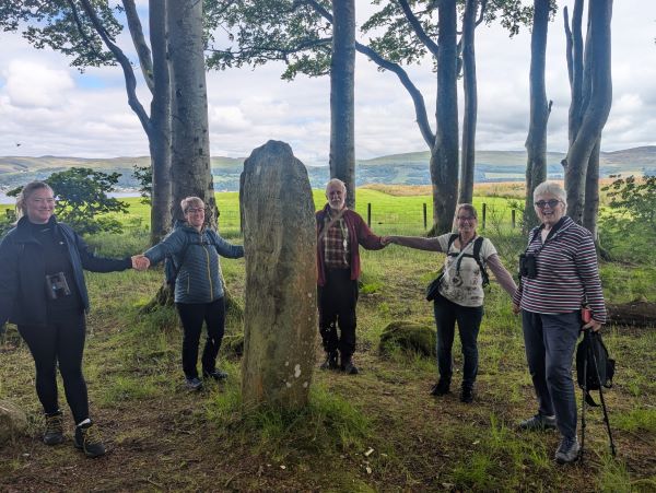 A group of people holding hands standing in a ring around a tall narrow stone.