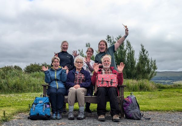 A group of people sitting on a bench with grass and trees in the background.