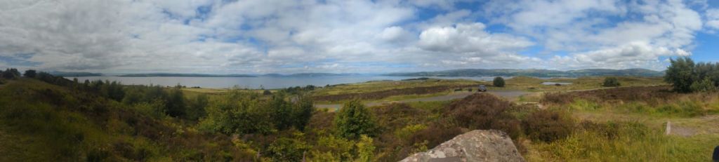 A panoramic landscape looking across grassland and scrub towards a body of water with land in the background.