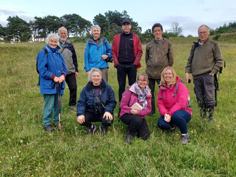 Group of 9 people wearing waterproofs, posing formally in a grassy field with trees in the background