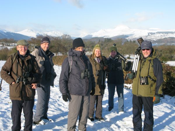 Six people wearing binoculars, waterproof and woolly hats standing in the snow with mountains behind them