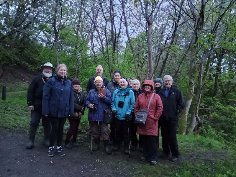 Group of people standing in Woodland at low light wearing waterproofs and binoculars