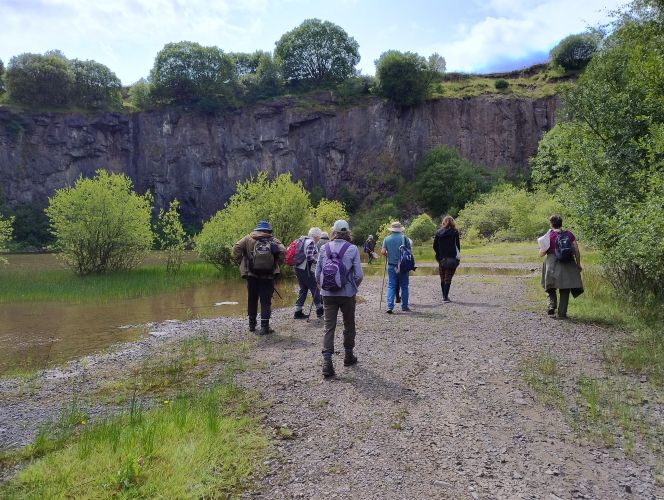 A group of people walking through a quarry along a gravelly path between trees and water 
