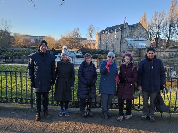 Group of six people standing in a row with an old building and waterfall in the background